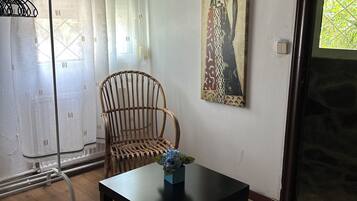 Une pièce avec un plancher en bois, une table noire sur laquelle est posé un vase bleu, une chaise en osier, un rideau blanc et un tableau accroché au mur.