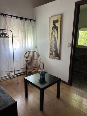 A room with a wooden floor, a black table with a blue vase, a wicker chair, a white curtain, and a painting on the wall.