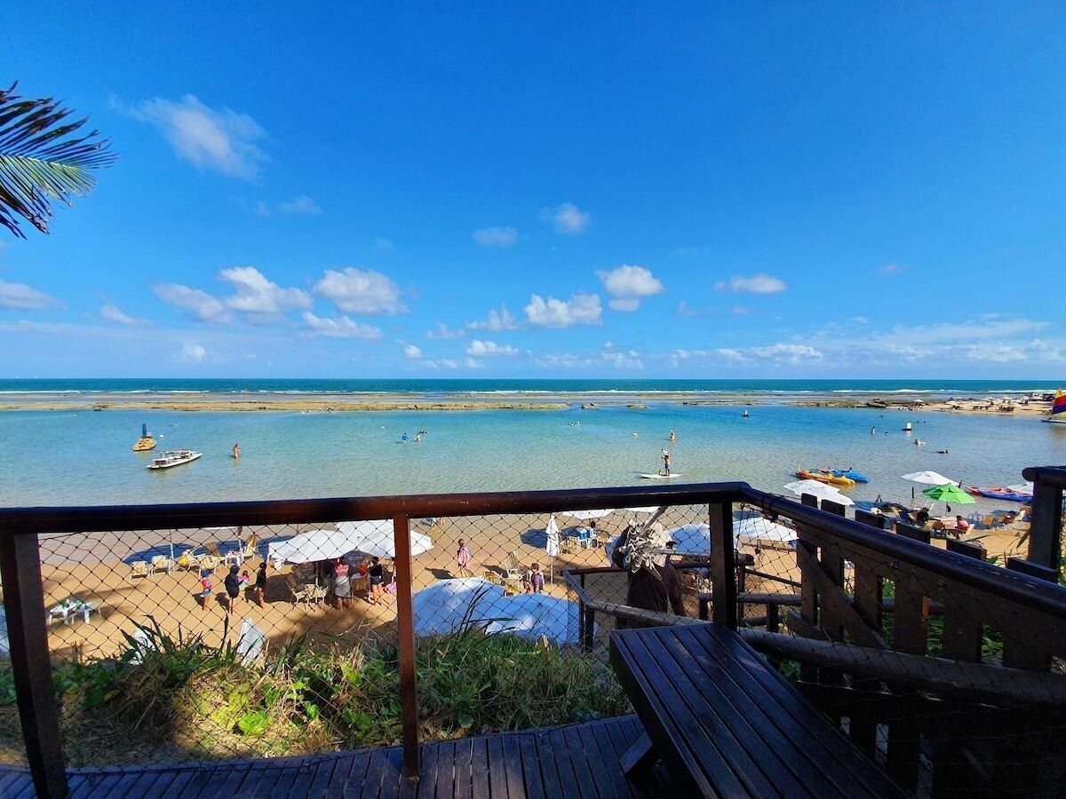 Una playa con muelle de madera, vista al agua, sombrillas y gente disfrutando.