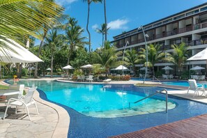 Una piscina con sillas reclinables y sombrillas, rodeada de palmeras y un edificio de varios pisos.