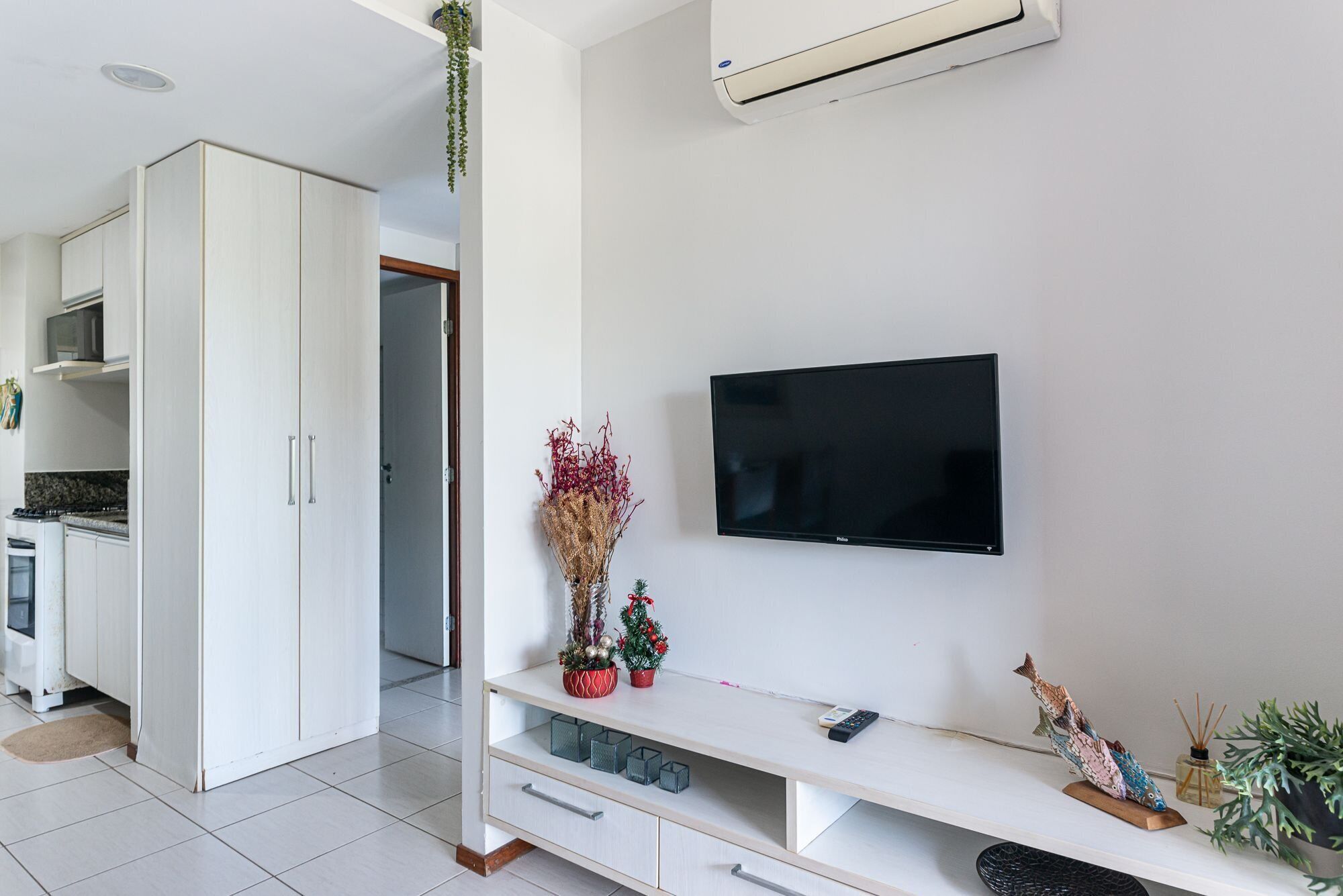 A modern living room with a flat-screen TV mounted on the wall, a white entertainment unit with drawers, and a kitchen area visible in the background.