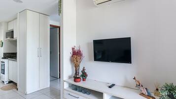 A modern living room with a flat-screen TV mounted on the wall, a white entertainment unit with drawers, and a kitchen area visible in the background.
