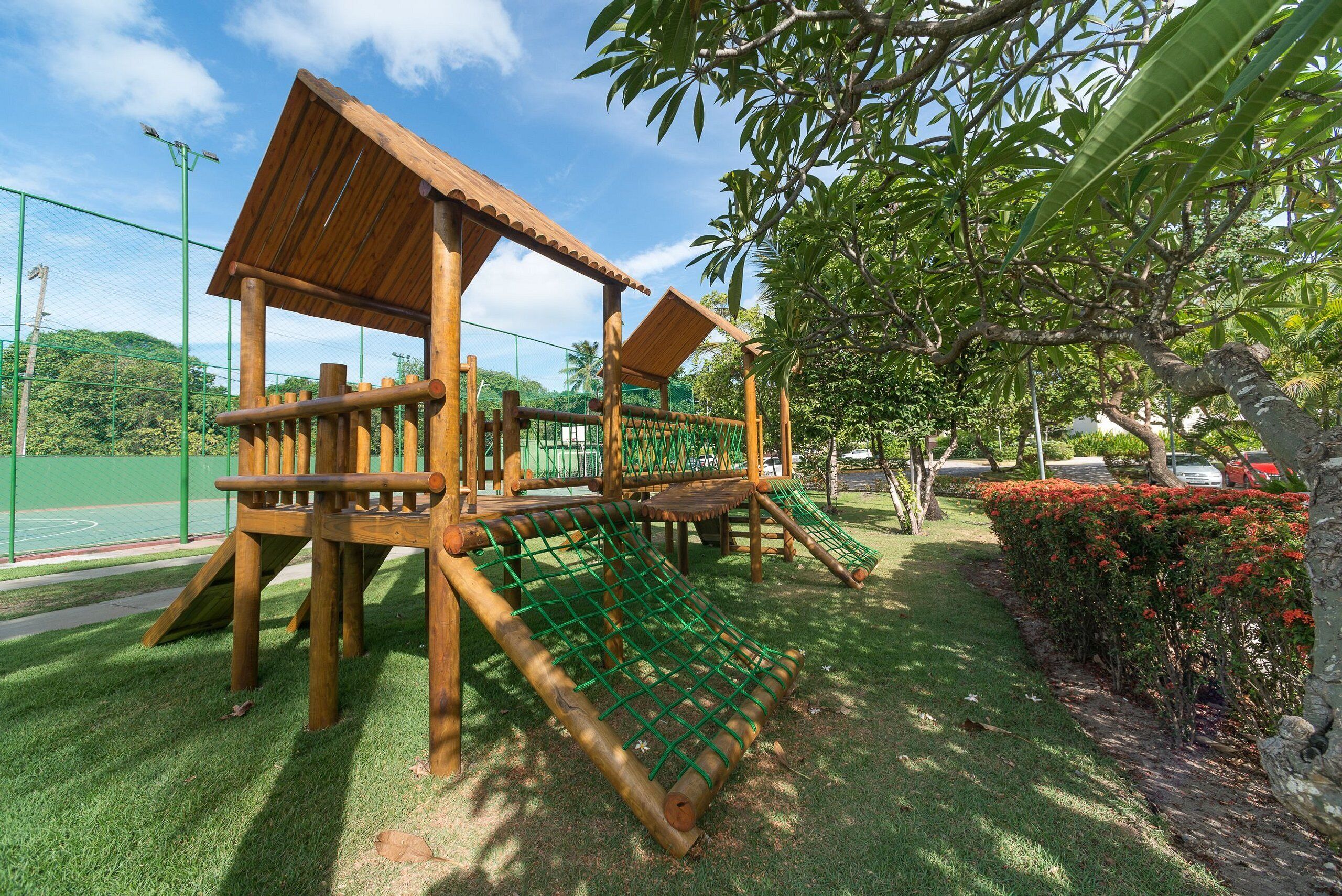 A playground with wooden structures, including a slide and climbing elements, surrounded by grass and trees.