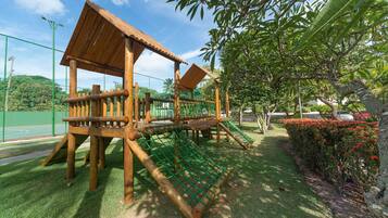 A playground with wooden structures, including a slide and climbing elements, surrounded by grass and trees.