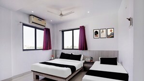 A hotel room with two beds, a ceiling fan, an air conditioning unit, a window with curtains, and a wall with three framed pictures.