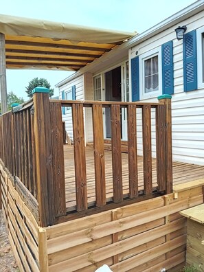 Eine Holzterrasse mit einem überdachten Pergola, ein weißes Gebäude mit blauen Fensterläden und ein klarer Himmel.