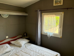 Ein Schlafzimmer mit einem Bett, einem Nachttisch, einer Lampe, einem Fenster mit Vorhang und einem an der Wand hängenden Bild.