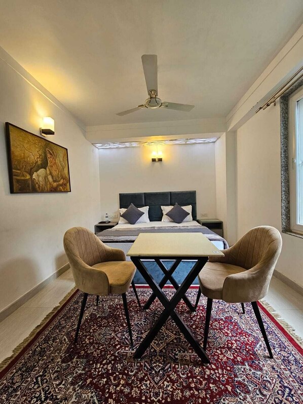 A hotel room with a bed, two armchairs, a table, a ceiling fan, a painting on the wall, and a patterned rug. - Sukon House by Crimson Horizon (Mussoorie)