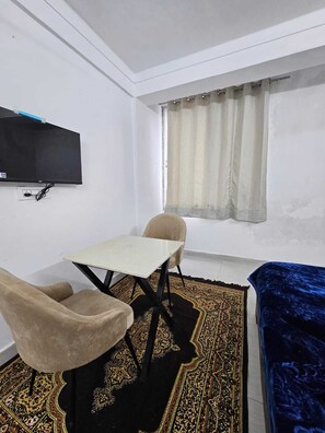 Una habitación pequeña y amoblada con televisión montada en la pared, una mesa, dos sillas, un sofá y una alfombra estampada.