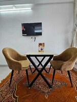 Una habitación con una televisión instalada en la pared, una mesita con un cartel, dos sillas y una alfombra estampada.