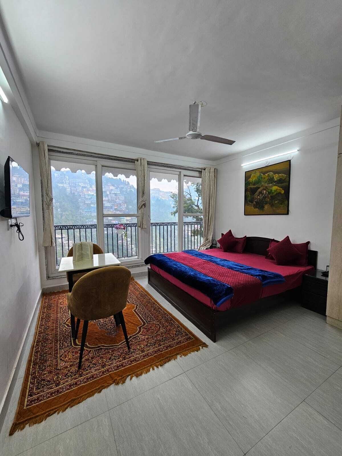 Un dormitorio con cama, una silla, una mesa, un cuadro y vistas de un paisaje urbano a través de amplios ventanales.