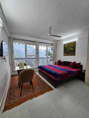 Un dormitorio con cama, una silla, una mesa, un cuadro y vistas de un paisaje urbano a través de amplios ventanales.