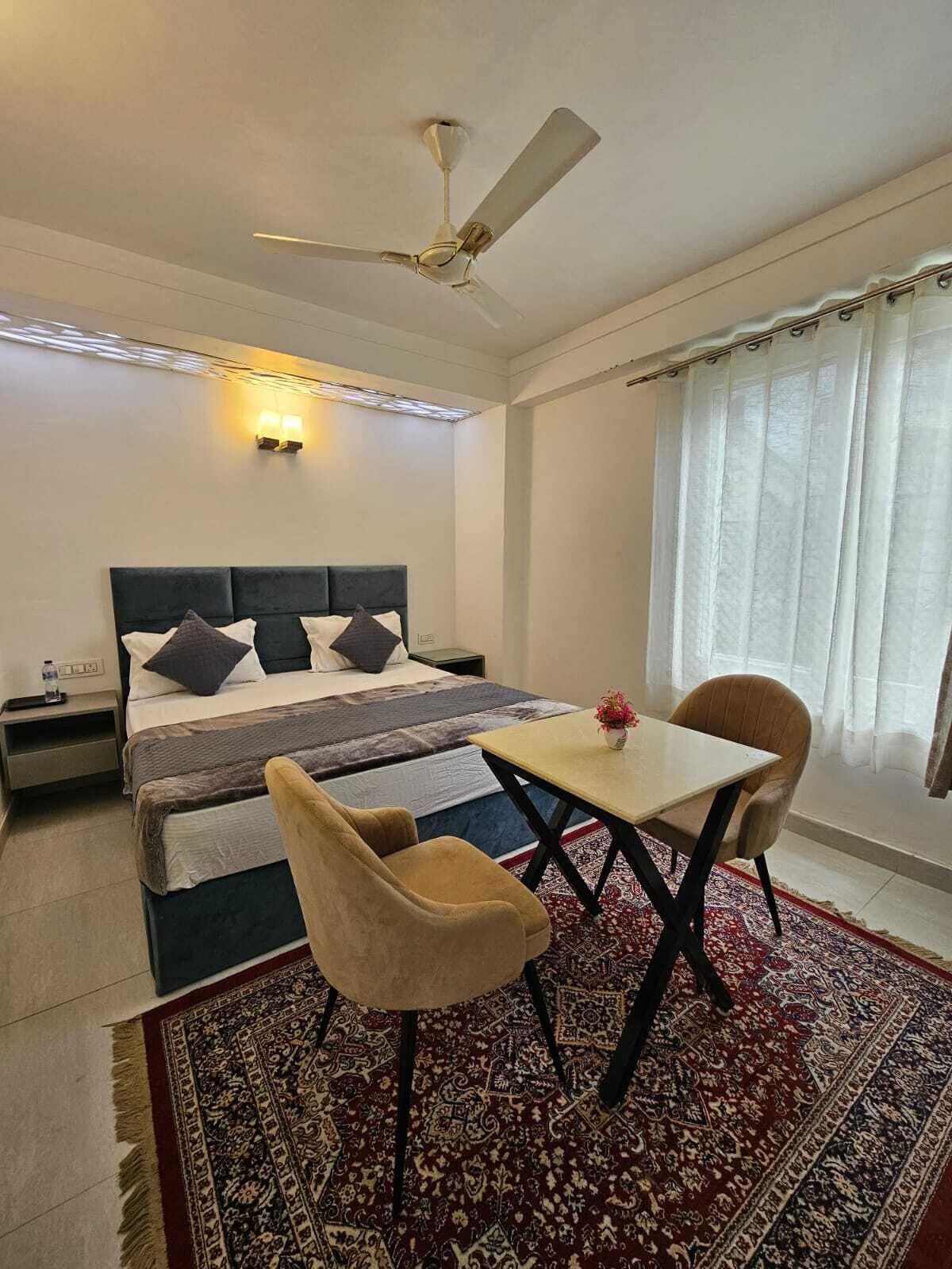 Een slaapkamer met een bed, nachtkastje, vloerkleed en een raam met gordijnen. Ook een tafel met twee stoelen en een plafondventilator.