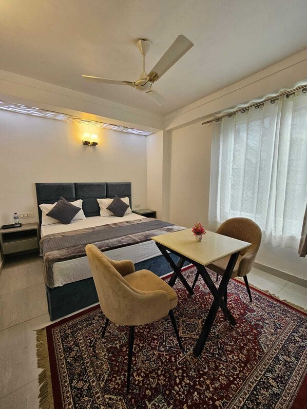 A bedroom with a bed, bedside table, nightstand, a ceiling fan, a table with two chairs, a rug, and a window with curtains.