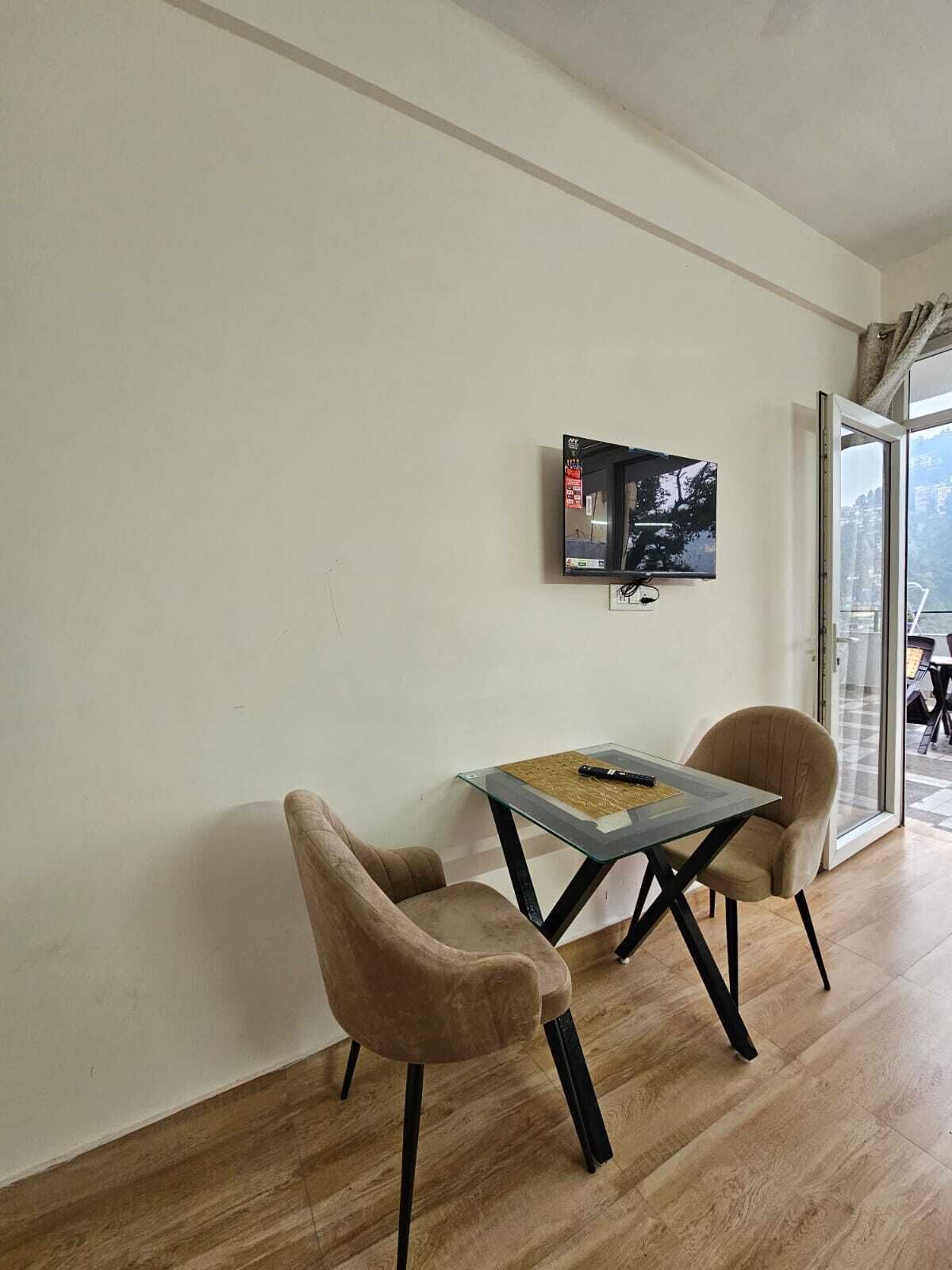 Een kamer met een glazen tafel en twee stoelen, een aan de muur bevestigde televisie en uitzicht door een glazen deur.