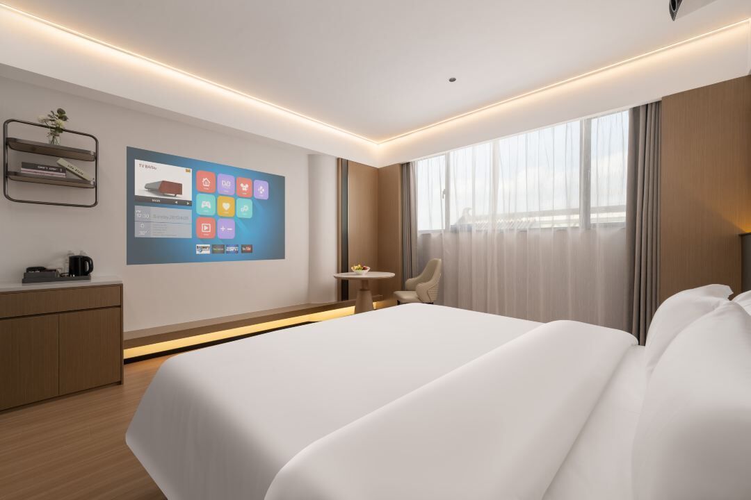 A modern hotel room with a large bed, a desk with a coffee maker, a small table with fruit, a chair, and a large window with curtains.