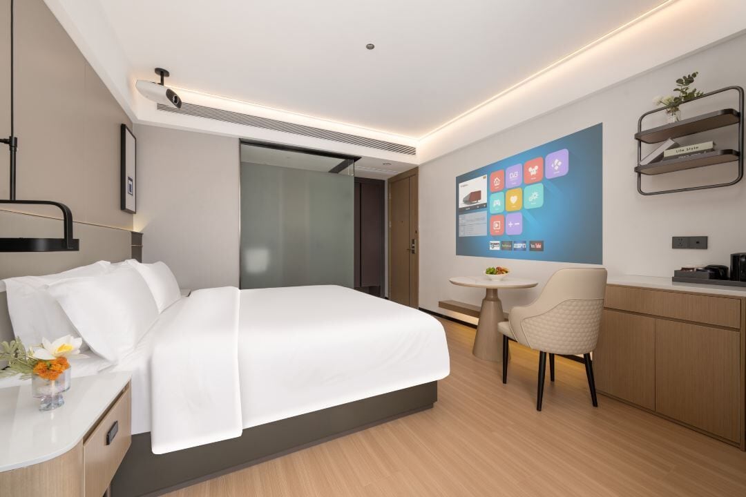 Una moderna camera d'albergo con un letto, una scrivania, una sedia, un tavolino e un display digitale a parete.