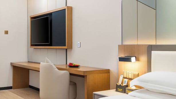 A hotel room with a wooden desk, a chair, a bed with white bedding, a bedside table with a lamp, and a small fruit bowl. - Poltton International Service Apartment (Foshan Yingtian Plaza Wanhua Metro Station (Foshan)
