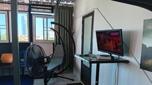 Una stanza con una scrivania, un monitor del computer, una ventola e una finestra vista edifici.