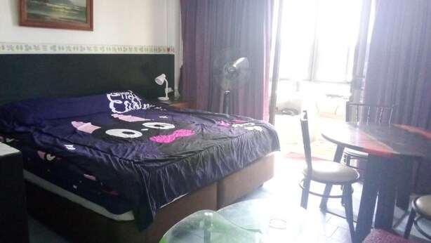 A hotel room with a bed, a small table, a chair, a fan, and a view of the outside. - Room-cozy Deluxe Guest Room in South Pat (Pattaya)