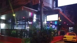 A nighttime view of a building with a large illuminated sign, a red barrier, and a yellow car parked outside. - Room-cozy Deluxe Guest Room in South Pat (Pattaya)