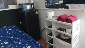 Ein kleines Zimmer mit einem Bett, einem kleinen Kühlschrank, einem weißen Schrank und einem Wandventilator.