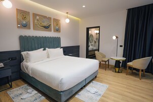 Habitación de hotel con una cama grande, una mesita de noche, una silla, una pequeña mesa y una obra de arte en la pared.
