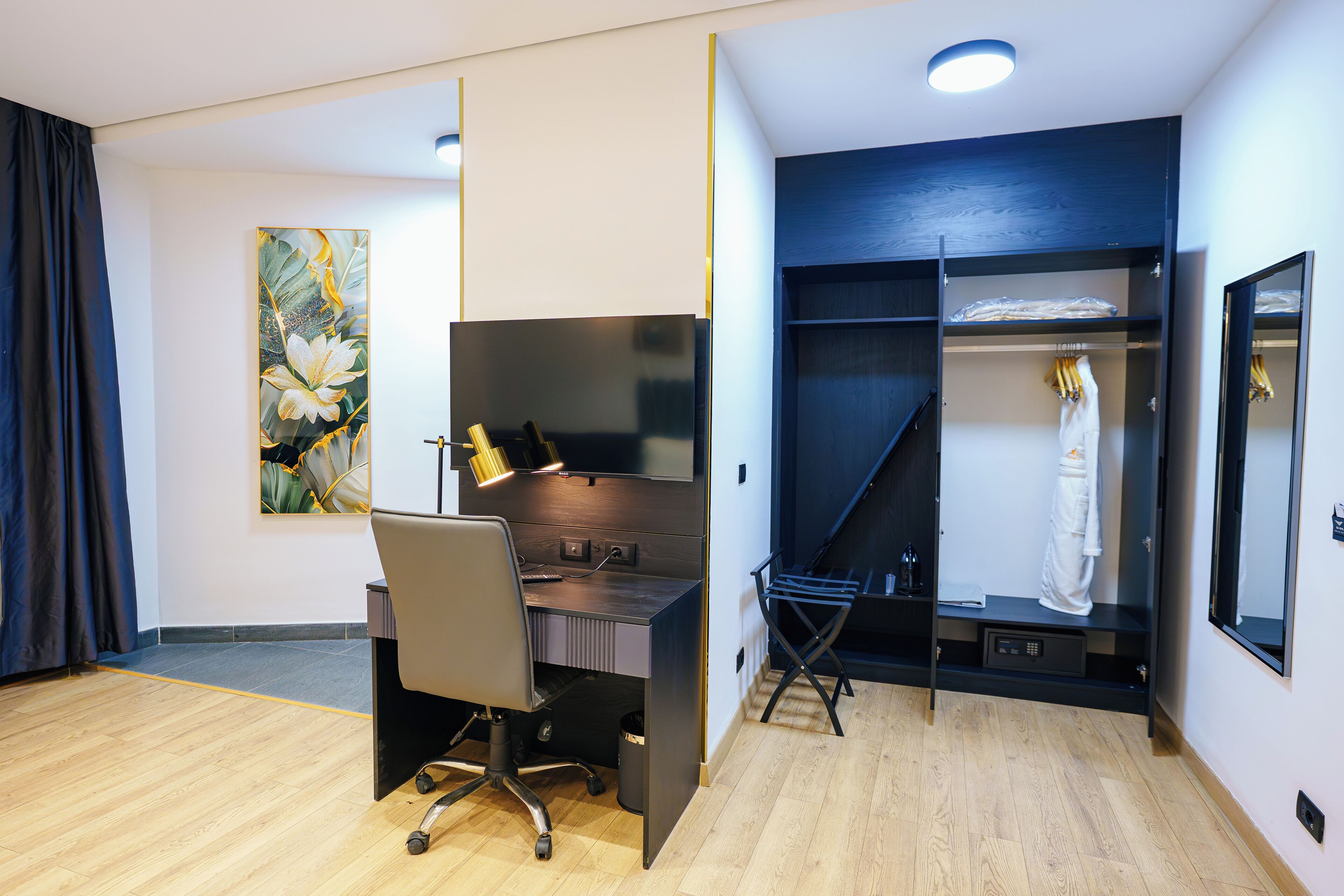 A hotel room with a desk, chair, wardrobe, and a painting on the wall.