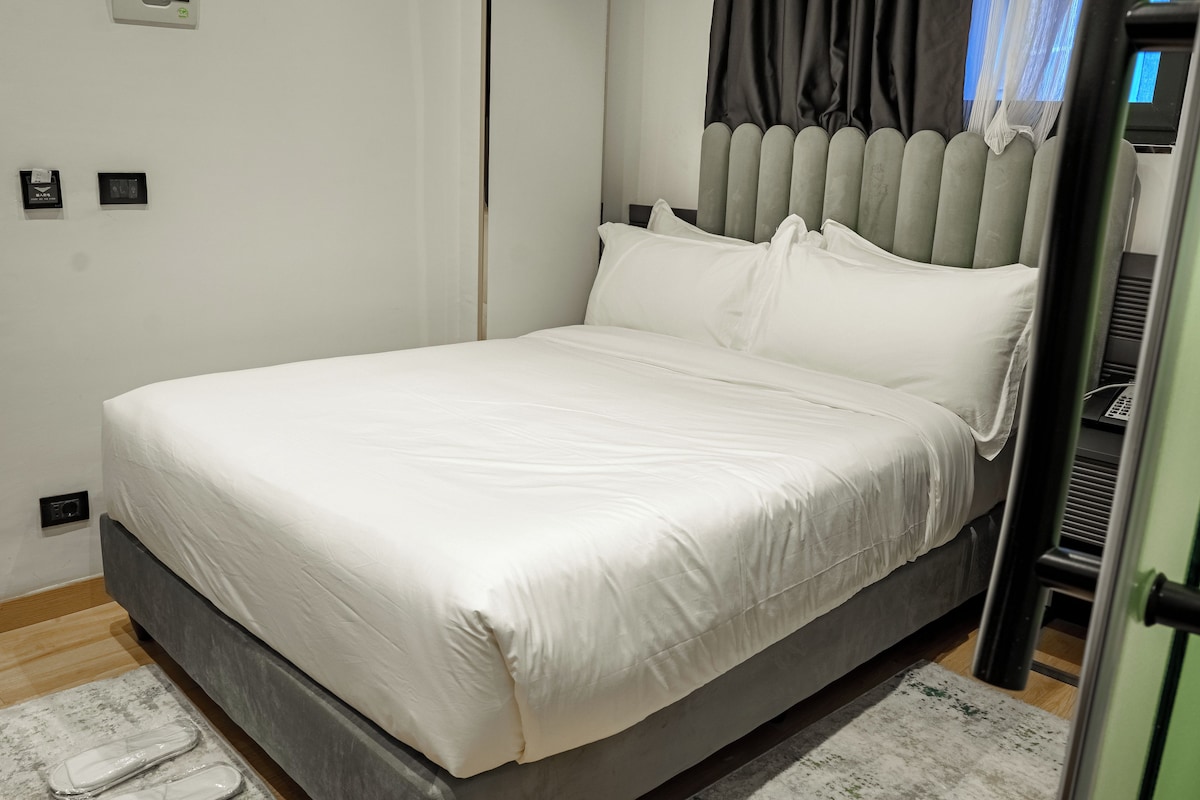 A neatly made bed with a grey headboard and white bedding in a modern room with a wooden floor and a small rug.