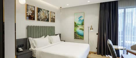 A modern bedroom with a large bed, a desk, and a chair. There are three paintings on the wall and a window with curtains.