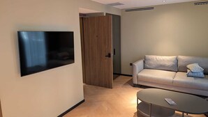 A modern living room with a flat-screen TV mounted on the wall, a grey sofa with cushions, a round coffee table, and a wooden door leading to another room. - Tanyel Hotel (Istanbul)
