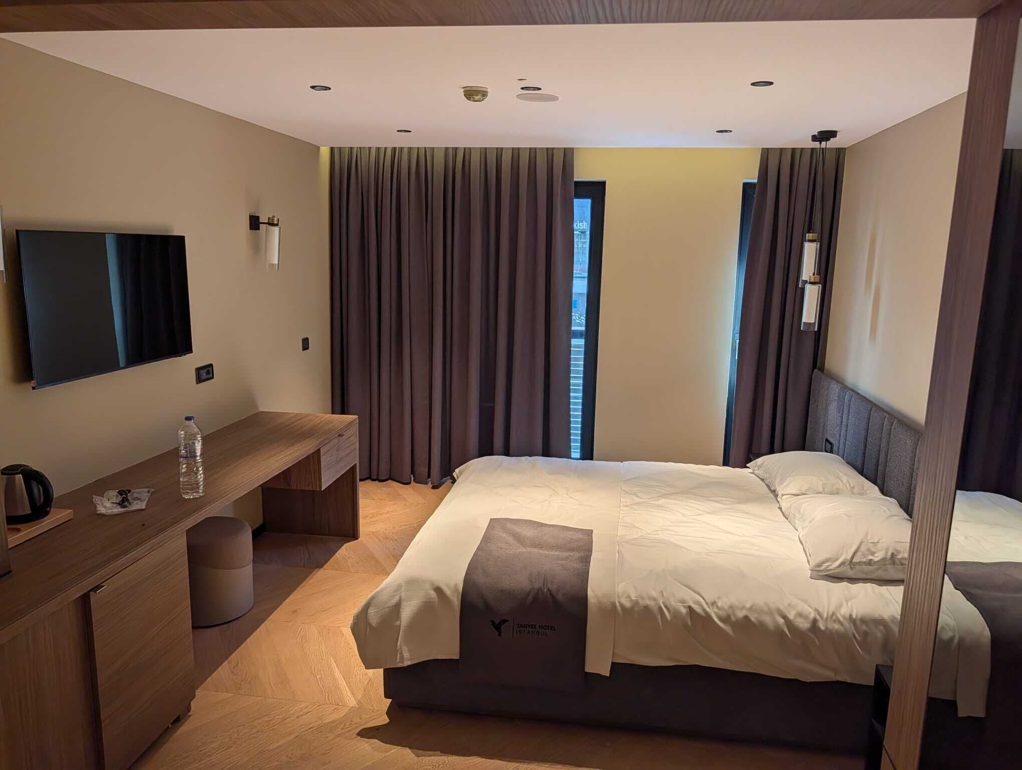 A hotel room with a bed, a desk with a chair, a television, and a window with curtains.
