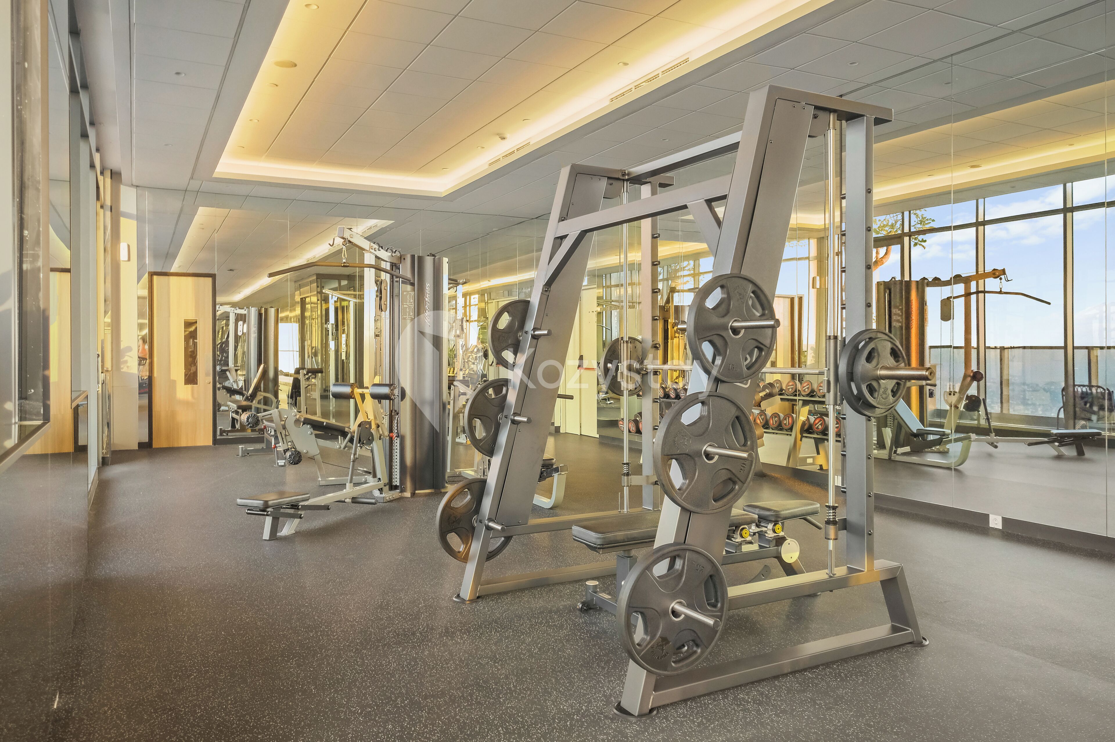 A gym with weightlifting equipment, mirrors, and large windows.