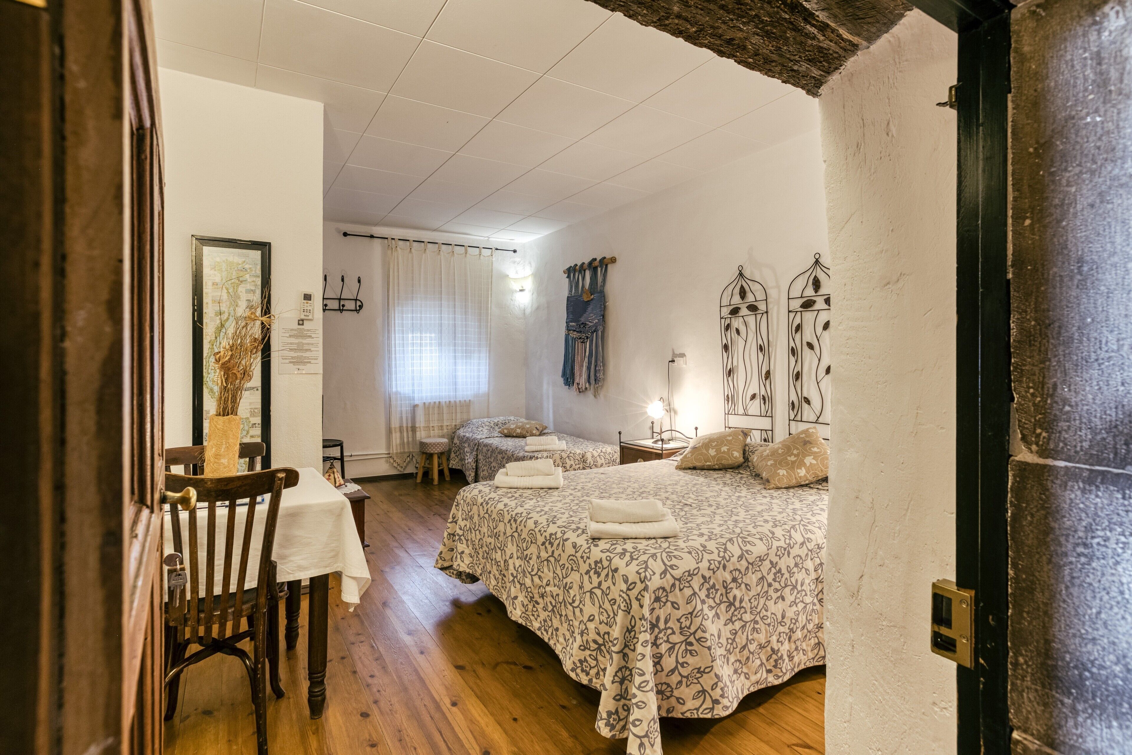Une chambre avec deux lits, un plancher en bois, un bureau avec une chaise, une fenêtre avec des rideaux et un luminaire fixé au mur.