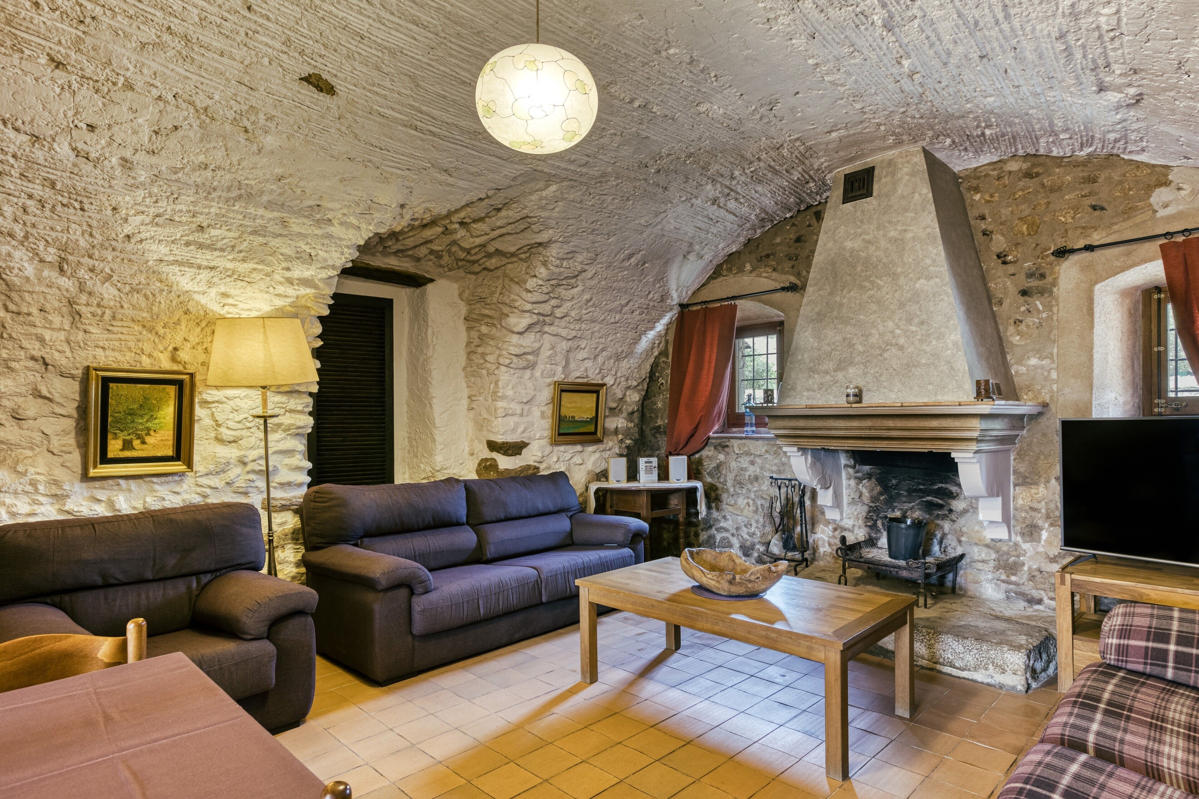Een rustiek interieur met een stenen plafond, een open haard, een bank, een televisie en een lamp.
