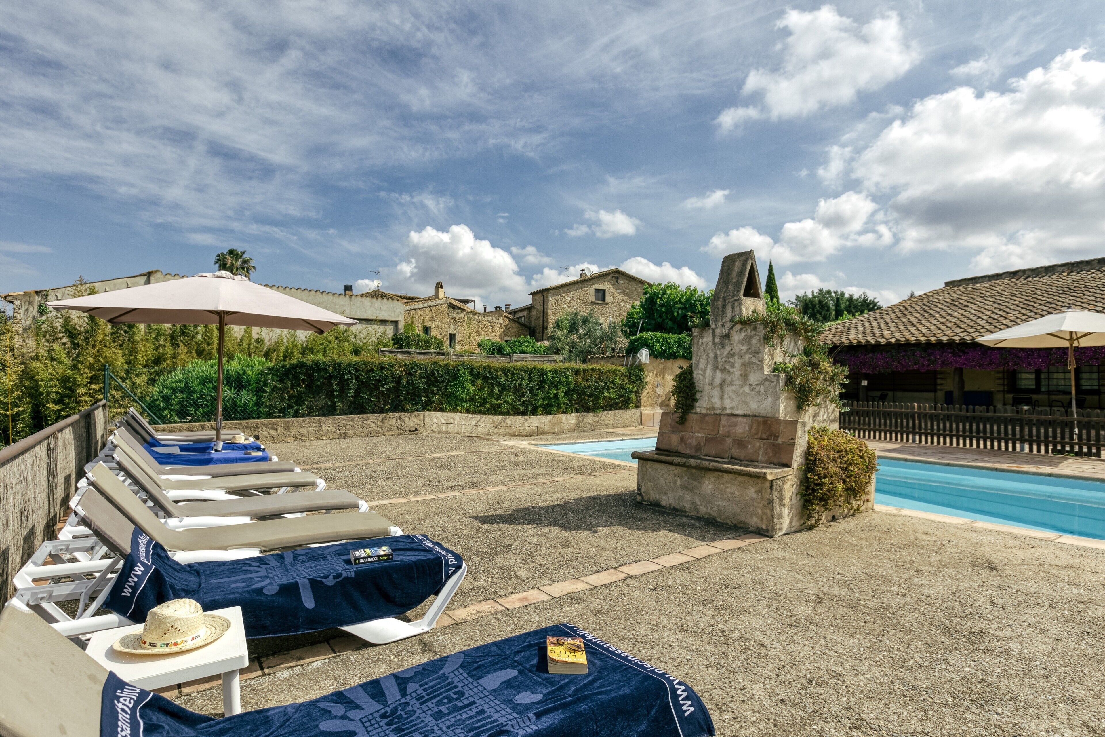 A poolside area with lounge chairs and umbrellas, a stone fountain, and a clear sky.
