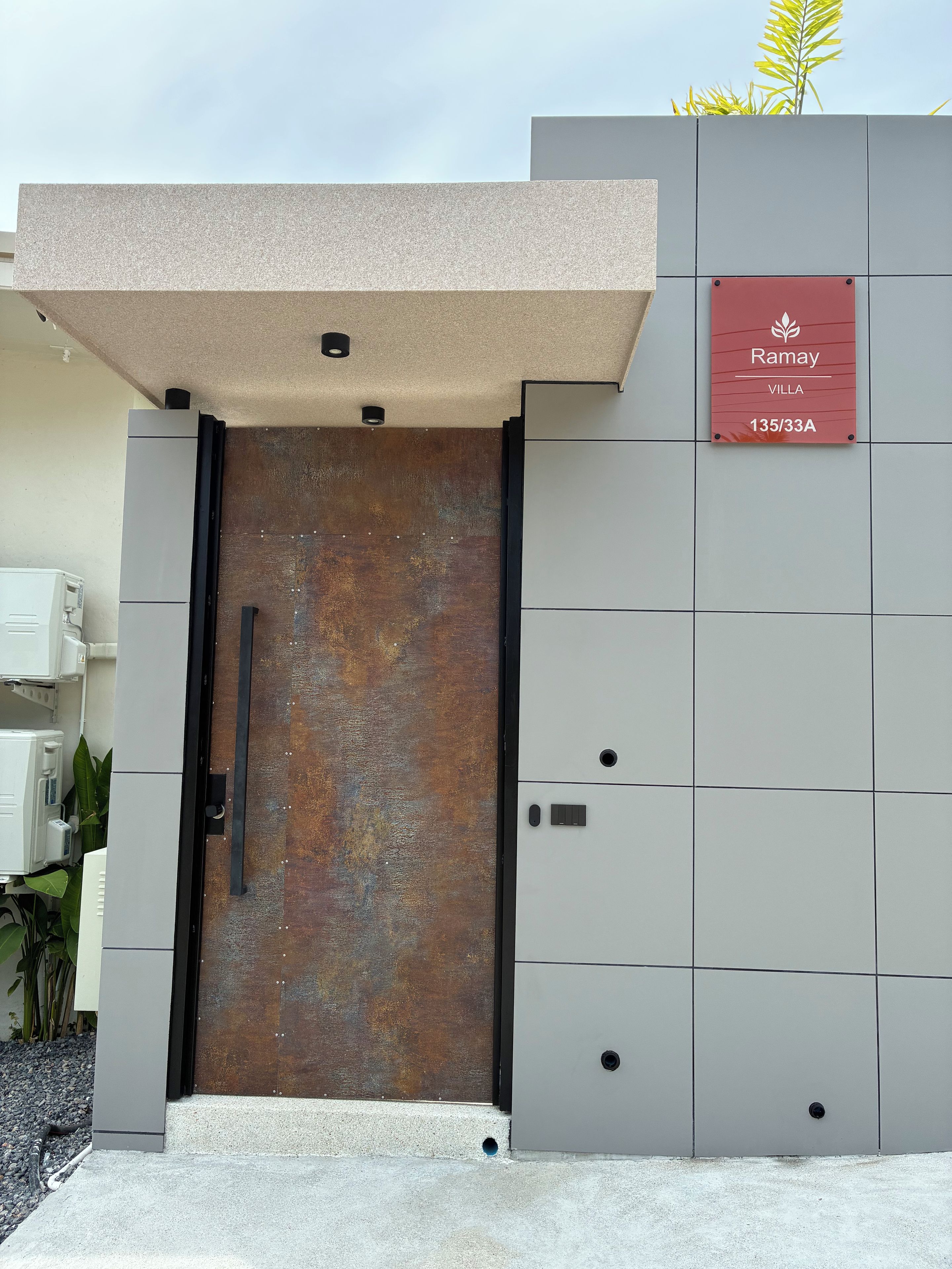 A gated entrance with a metal door, a sign indicating 'Ramay Villa 135/33A', and a small outdoor area with plants.