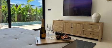 Un salon moderne doté d’une grande fenêtre, d’un téléviseur à écran plat, d’un meuble en bois, d’un coin salon confortable et d’un plateau sur lequel se trouvent une bouteille et des verres.