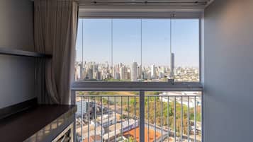 A room with a large window overlooking a cityscape, a balcony with a metal railing, and a dark-colored desk.