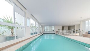 An indoor swimming pool with clear glass walls, a tiled floor, and a view of the cityscape. - Flat w/ gym in Setor Bueno - Wbu2405 (Goiânia)