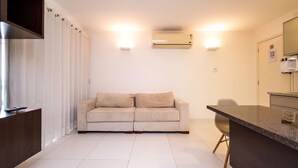 A modern living room with a beige sofa, a kitchen area with a countertop, and a wall-mounted air conditioner. - Flat w/ gym in Setor Bueno - Wbu2405 (Goiânia)