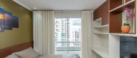 A hotel room with a bed, a TV, a balcony with curtains, and a city view.