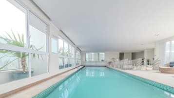 Una piscina interior con paredes de vidrio transparente, suelo de baldosas y una zona de estar con sillones de descanso.