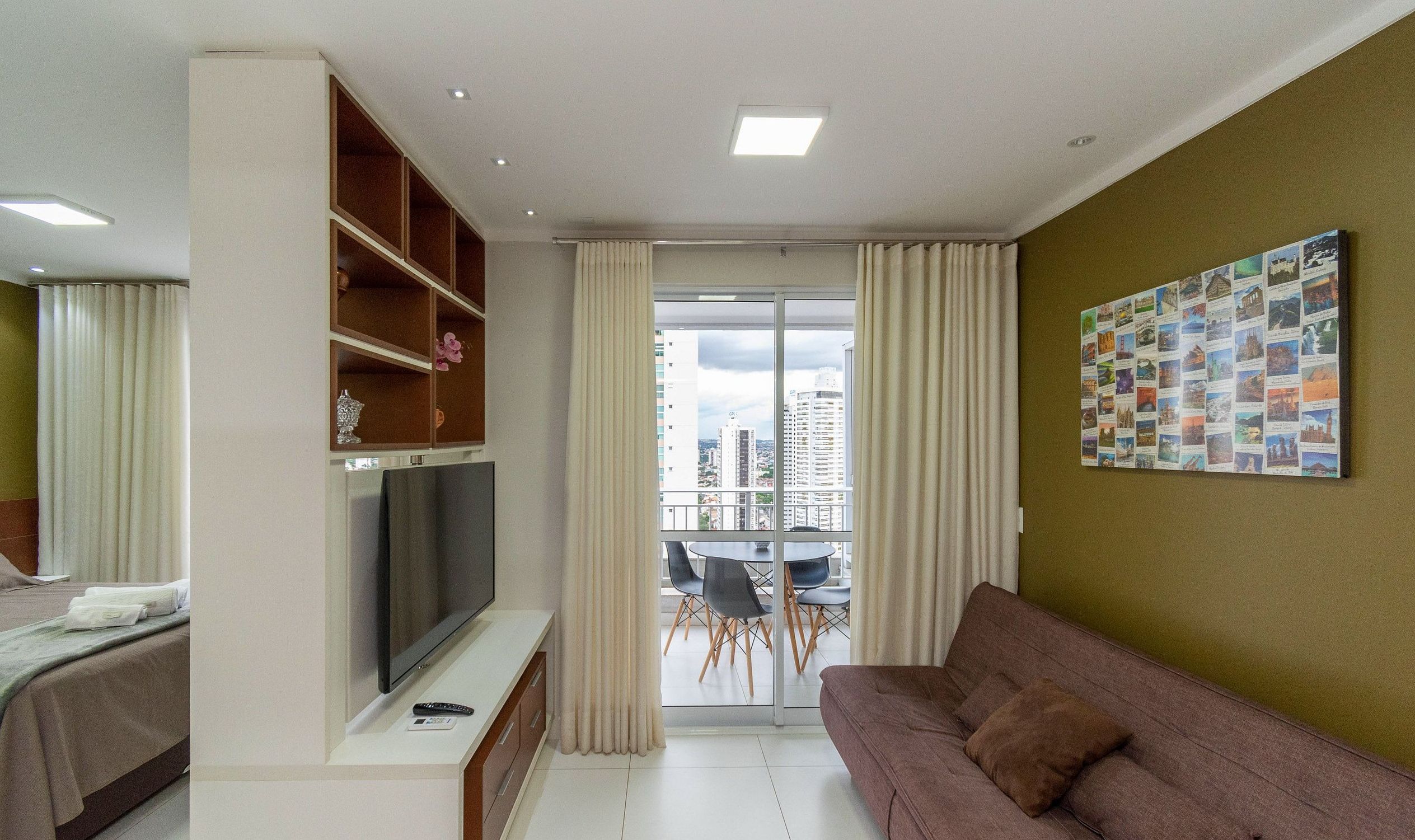 A modern living room with a flat-screen TV, a sofa, a dining area with chairs, and a city view through the glass door.