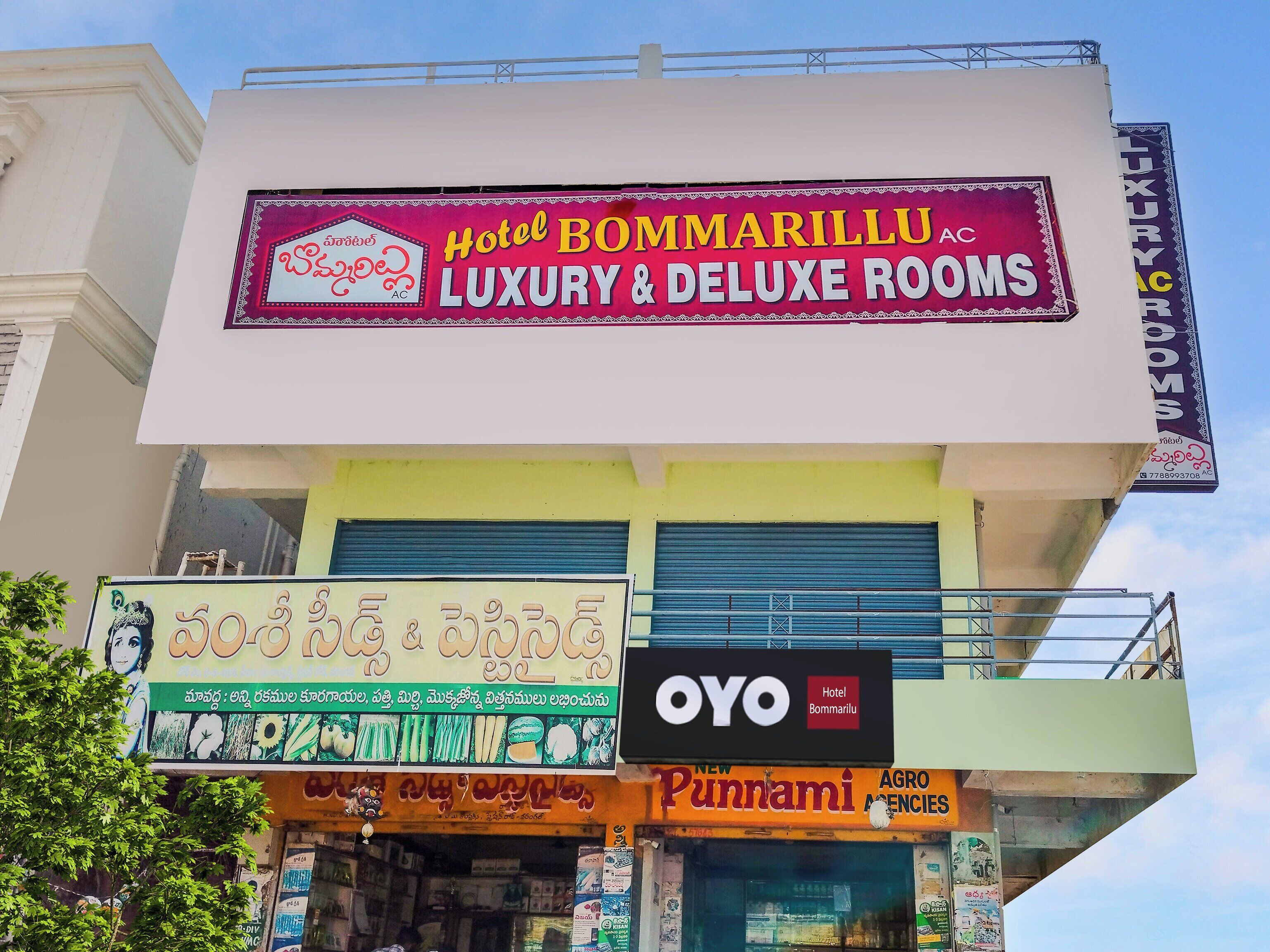 Un hotel llamado Hotel Bommarillu, con carteles que indican habitaciones de lujo y superiores, una agencia Punnami Agro, y un cartel de OYO Hotel.