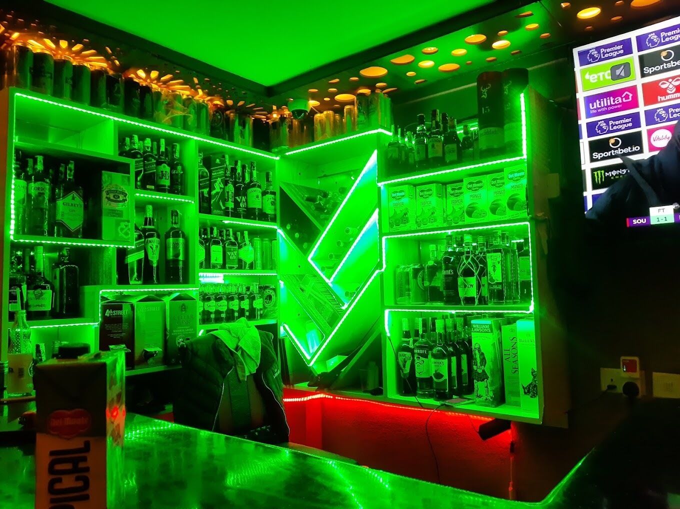 A bar area with shelves stocked with various liquor bottles, a neon green arrow sign, and a digital screen displaying sports betting logos.