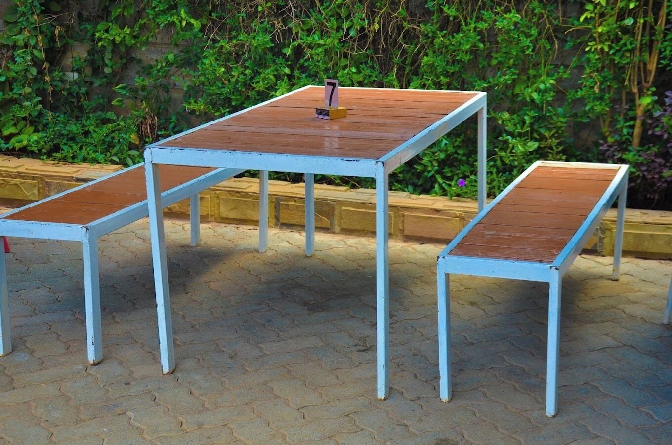 Un conjunto de tres mesas metálicas blancas con tapas de madera, numeradas 7, 8 y 9, ubicadas en un espacio al aire libre empedrado con un seto verde al fondo.