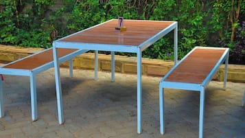 A set of three white metal tables with wooden tops, numbered 7, 8, and 9, placed on a paved outdoor area with a green hedge in the background.