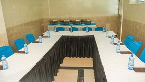 Un salón de conferencias con una mesa larga, sillas azules, botellas de agua y una cortina negra.