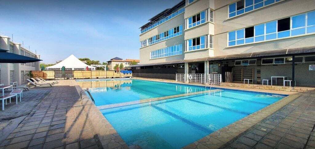 Una piscina con solárium empedrado, junto a un edificio con varias ventanas y una zona cubierta al fondo.